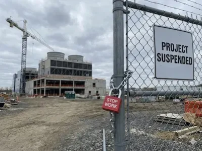 Baustelle mit unfertigem Industriegebäude hinter Zaun; Schild „Project Suspended“ und Schloss mit „Stop Work“.